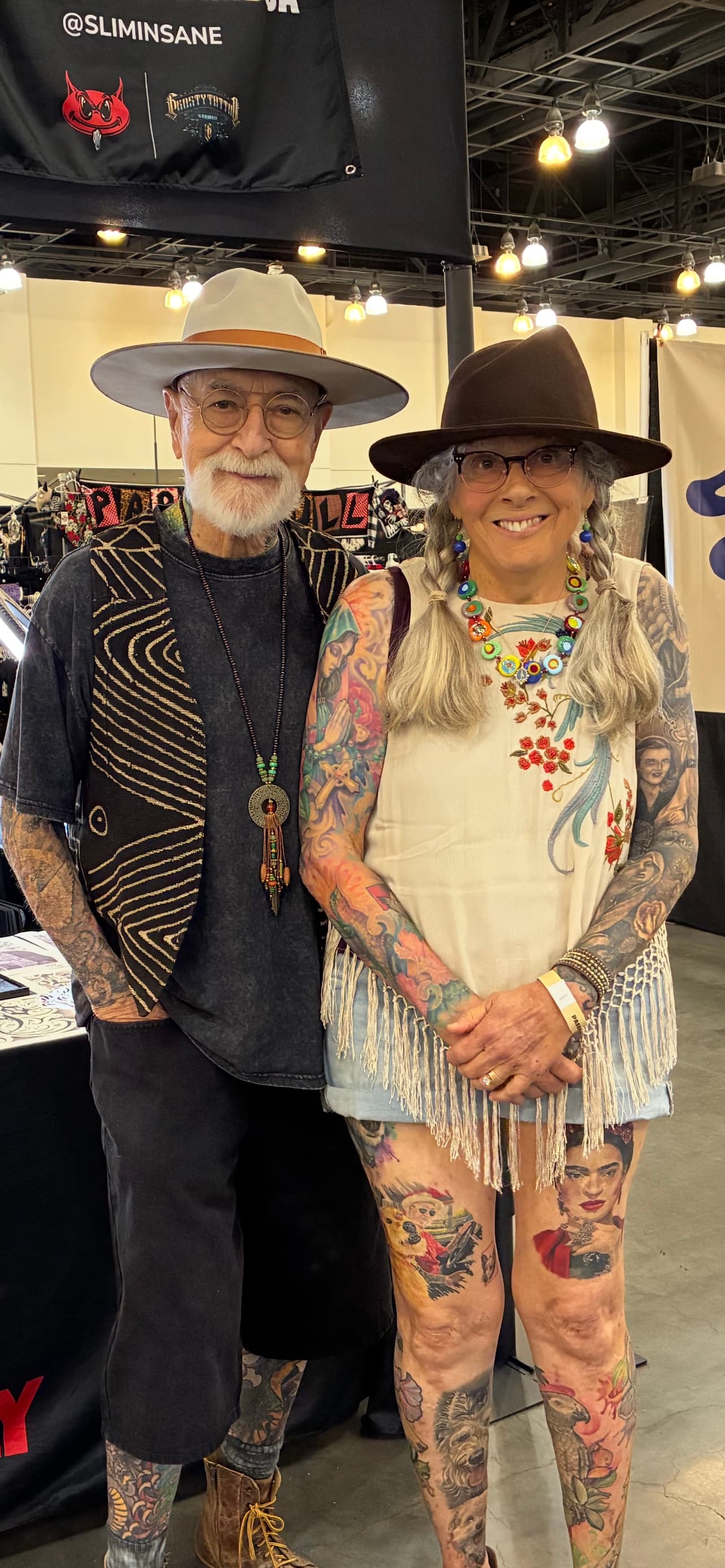 Bill and Ann smiling together in wide-brimmed hats and showing tattoos collected across their arms and legs