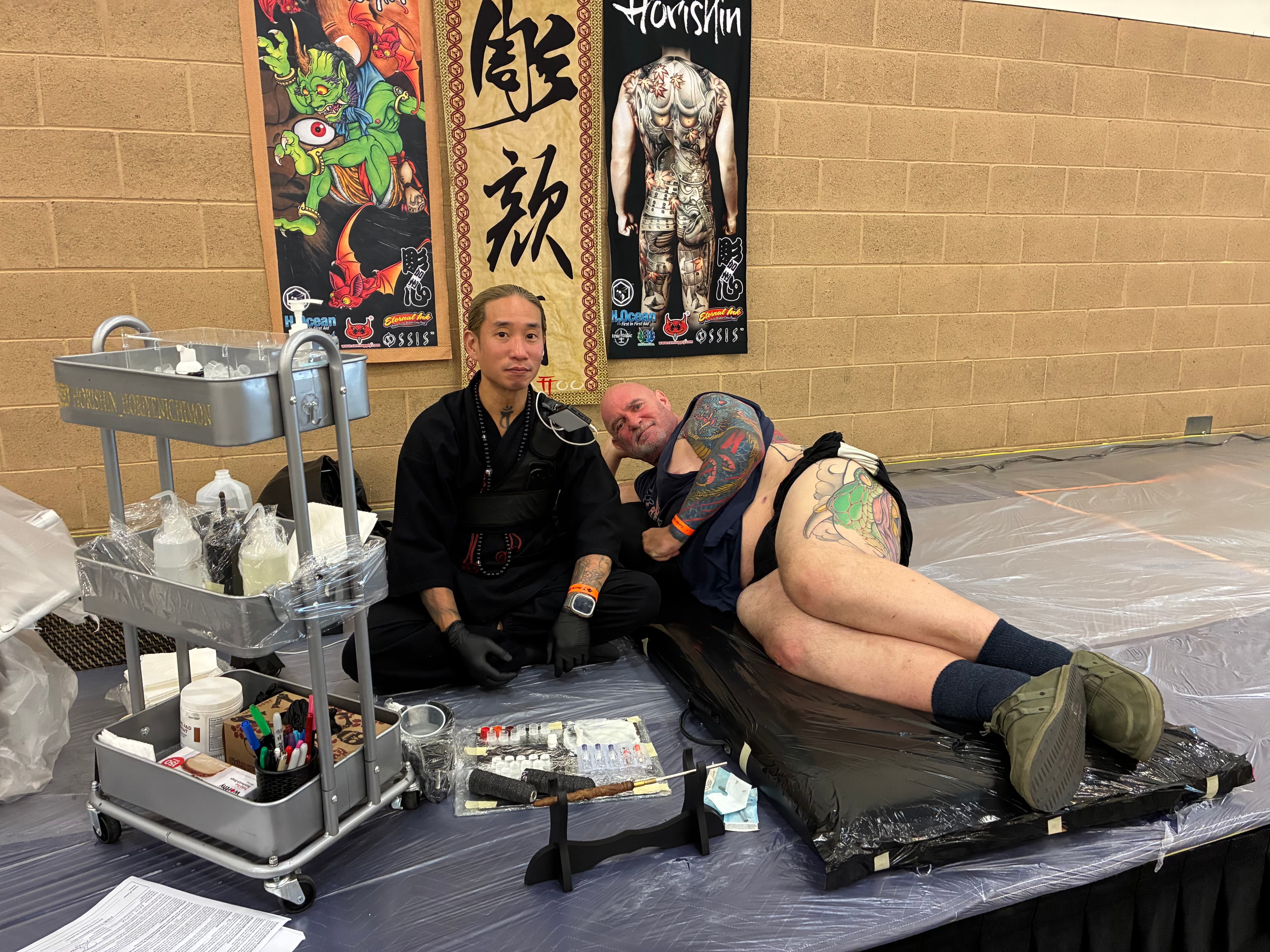 Bobby lying on a mattress beside his tattoo artist at the Golden State Tattoo Expo