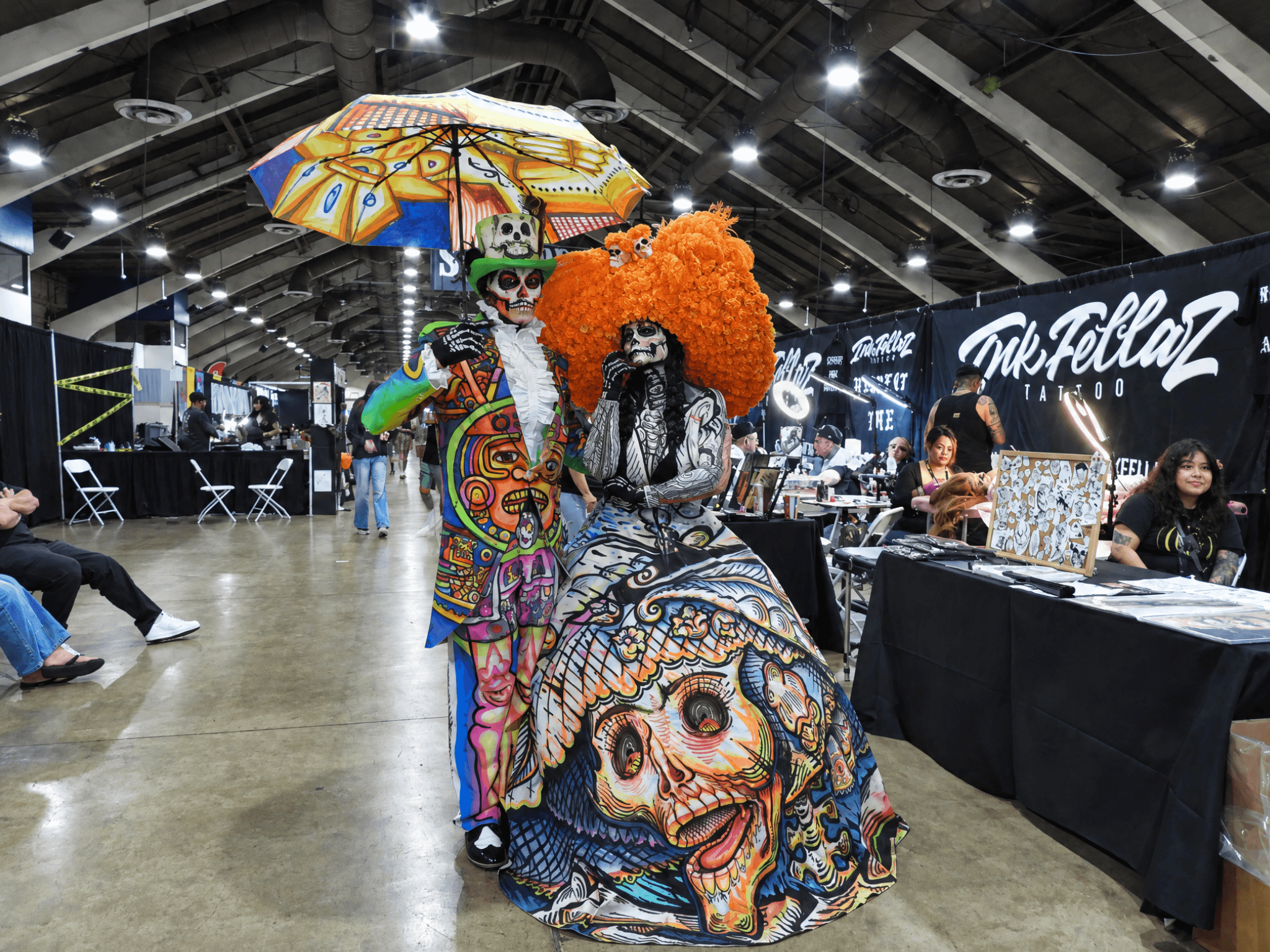 Maria Flores as La Catrina in an orange floral headdress and illustrated dress, beside a man in a colorful skull-painted suit holding a painted umbrella