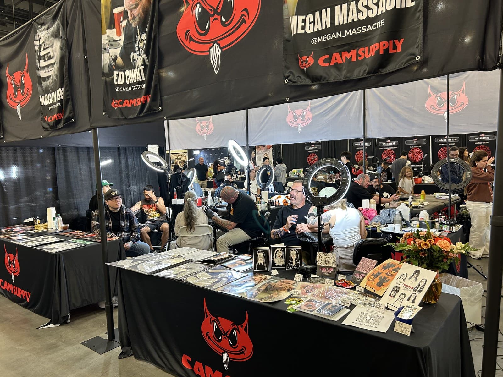 Busy tattoo convention floor viewed across artist booths, ring lights, tattooing stations and artwork displays