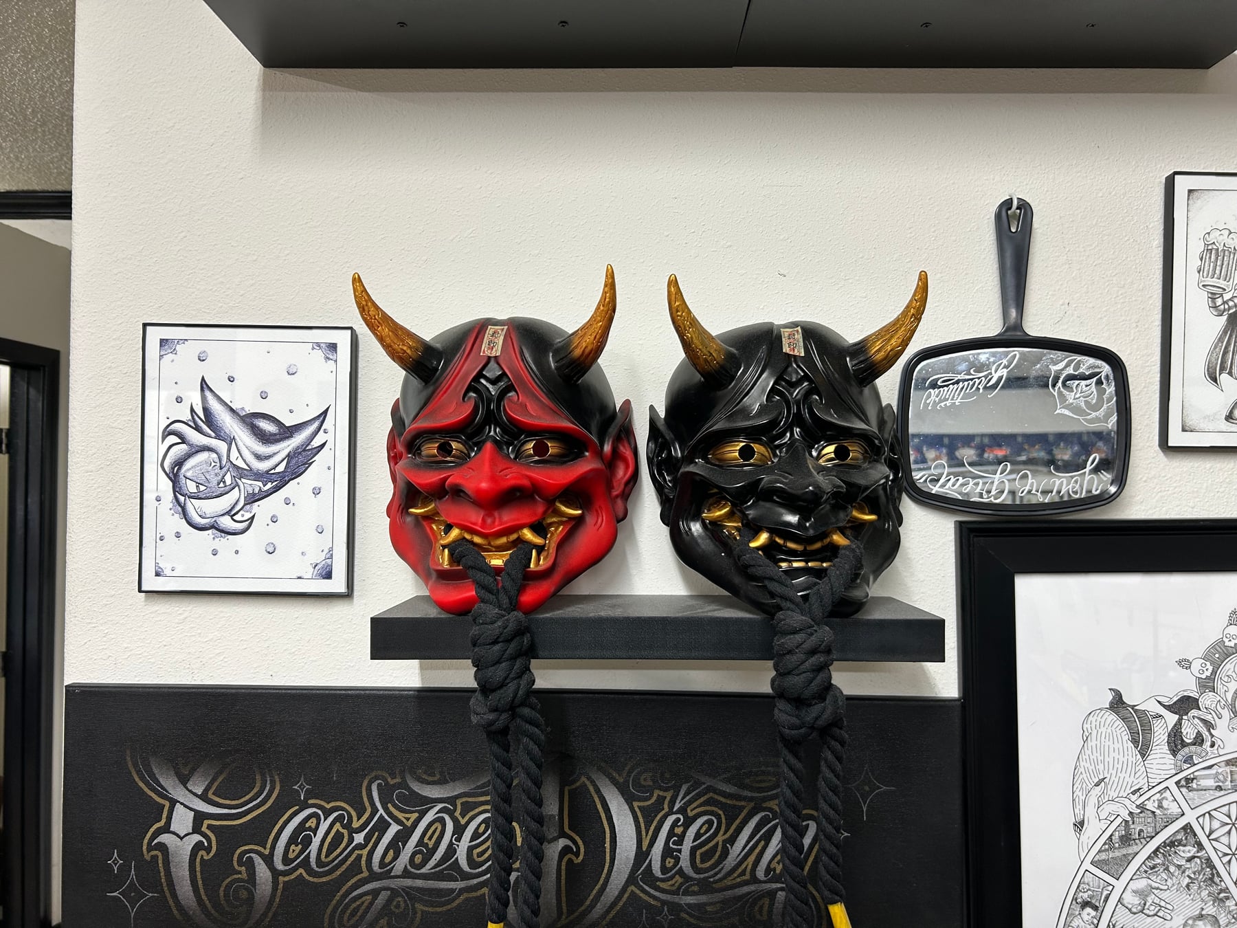 Red and black Japanese Oni masks displayed inside a tattoo shop beneath framed tattoo artwork